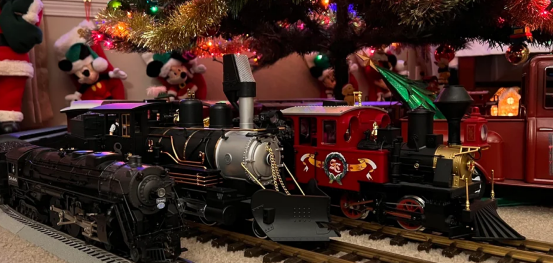 Toy trains circle under a decorated Christmas tree with colorful lights and tinsel. Plush Disney characters, including Mickey and Minnie Mouse, are visible in the background near holiday decorations.