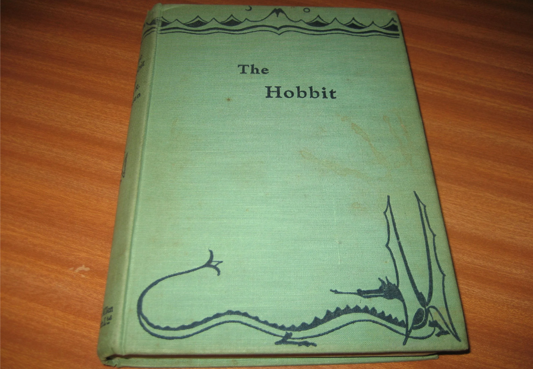 A worn green hardcover edition of "The Hobbit" lies on a wooden surface. The cover features black illustrations of a dragon and decorative borders, with the title centered in plain text.