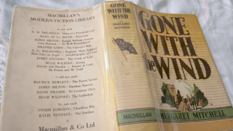 A hardcover copy of "Gone with the Wind" by Margaret Mitchell sits open facedown, showing both the yellow dust jacket with the title and author, and an inside page listing other books in Macmillan&rsquo;s Modern Fiction Library.