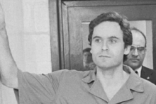 A man in a prison jumpsuit stands with his arm raised inside a building, while another man in a suit and glasses stands behind him. The scene appears to be indoors, possibly in a courthouse or police station.