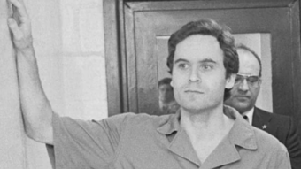 A man in a prison jumpsuit stands with his arm raised inside a building, while another man in a suit and glasses stands behind him. The scene appears to be indoors, possibly in a courthouse or police station.