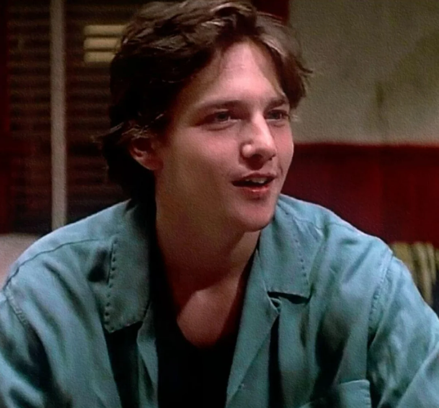 A young man with medium-length brown hair wears a teal jacket and smiles while looking slightly to the side, sitting indoors with a blurred background featuring wooden blinds and a red wall.
