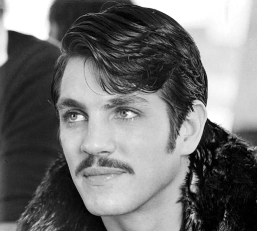 A black and white photo of a young man with neatly styled dark hair, a mustache, and light-colored eyes. He is wearing a fur-collared coat and looking slightly to the side with a neutral expression.