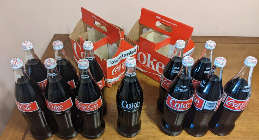 Twelve glass bottles of Coca-Cola and Coke, arranged in front of two vintage red and white cardboard bottle carriers, all placed on a wooden table against a beige wall.