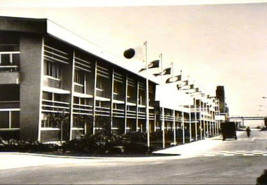 Black and white photo of a modernist two-story building with many windows and a row of flags lining the sidewalk. A few people and vehicles are visible near the entrance. The scene appears quiet and orderly.