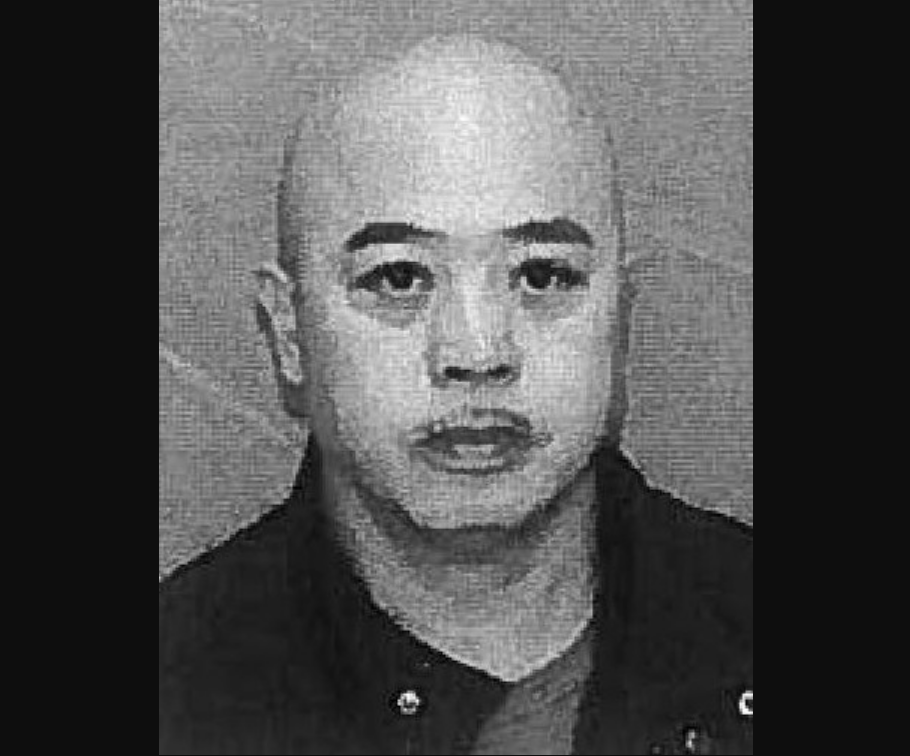 Black and white photo of a person with a bald head, dark eyebrows, and wearing a dark jacket over a light shirt, facing forward with a neutral expression.