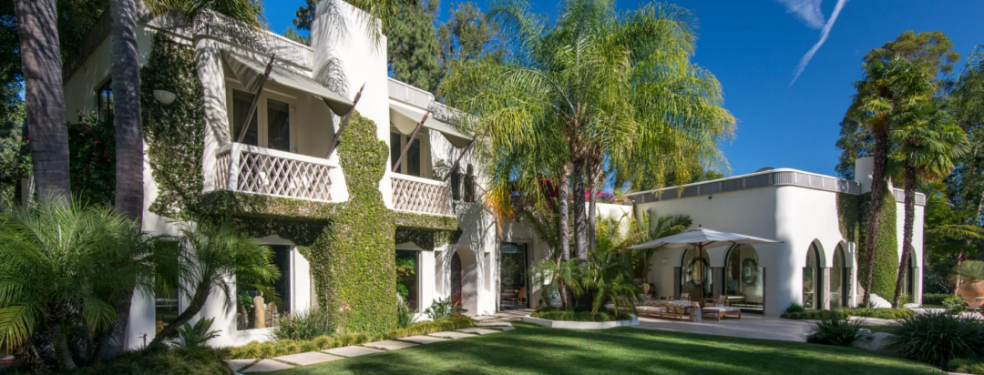 A modern white villa with ivy-covered walls, balconies, large arched windows, and a patio area with tables and umbrellas, surrounded by lush palm trees and greenery under a clear blue sky.