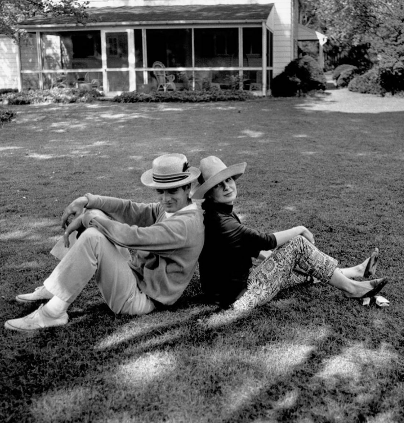 Two people wearing hats sit back-to-back on a grassy lawn, both looking toward the camera. A house with a porch is visible in the background, surrounded by trees and garden.