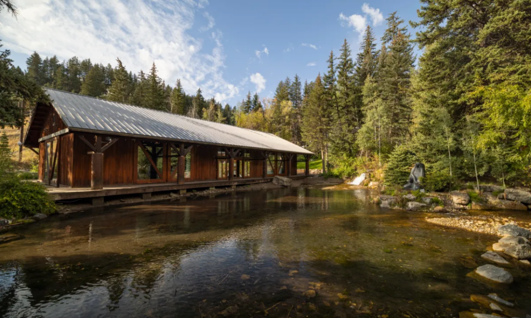 A wooden covered bridge stands over a clear, shallow stream surrounded by tall pine trees and dense forest under a partly cloudy sky. Sunlight illuminates the scene, creating reflections in the water.