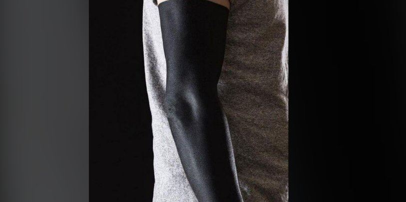 A person wearing a gray T-shirt and black shorts has their arm extended, showing a solid black tattoo sleeve covering the entire arm and a geometric starburst pattern tattooed on the hand.