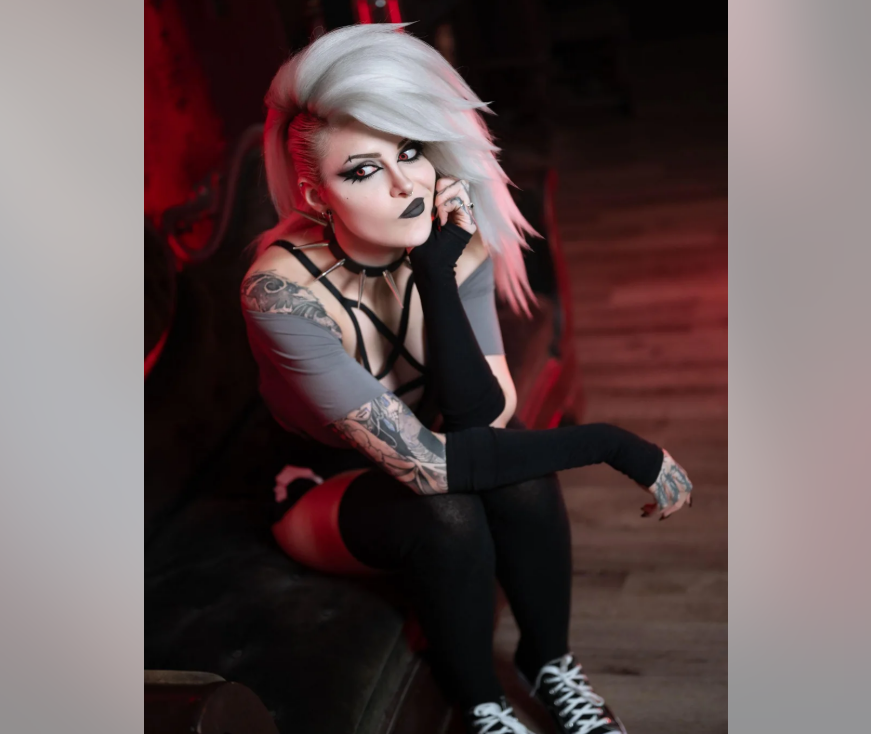 A person with striking white, voluminous hair and dramatic black makeup sits on a vintage sofa, wearing a grey top, black crisscross harness, arm warmers, and thigh-high socks. Tattoos are visible on their arms. The lighting is moody.