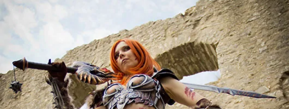 A person in elaborate fantasy armor with red hair holds a sword and mace, standing in front of ancient stone ruins under a partly cloudy sky.