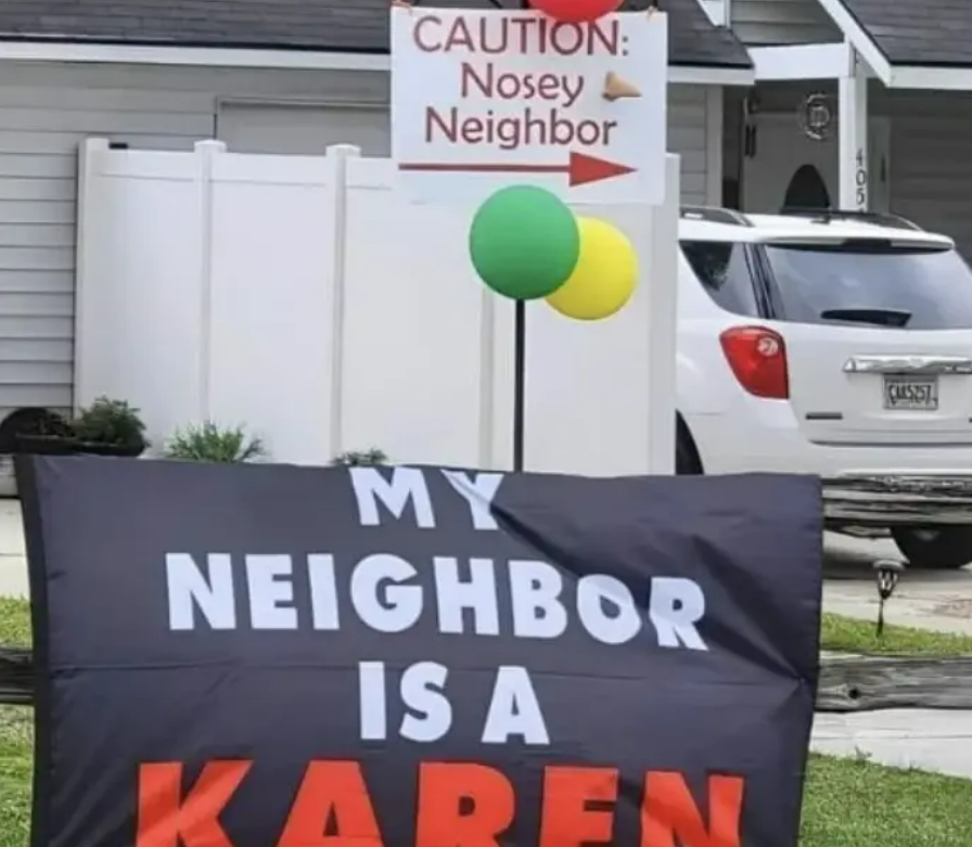 A yard sign reads "MY NEIGHBOR IS A KAREN" in bold letters. Behind it, another sign with an arrow says "CAUTION: Nosey Neighbor." A white car and a house are visible in the background.