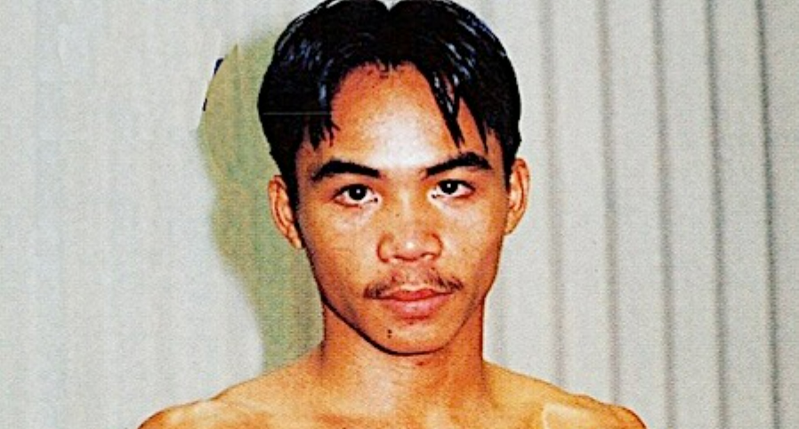 A young man with short dark hair and a mustache stands shirtless, holding his fists up in a boxing stance in front of a plain, vertical-striped background.