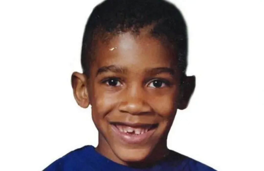 A young boy with short hair smiles widely, showing a gap between his front teeth. He is wearing a blue shirt and is pictured against a plain white background.