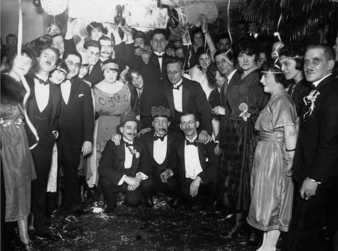A large group of people dressed in formal 1920s attire celebrate at a festive indoor party, with streamers hanging and confetti on the floor. Most are smiling and posing closely together for the camera.