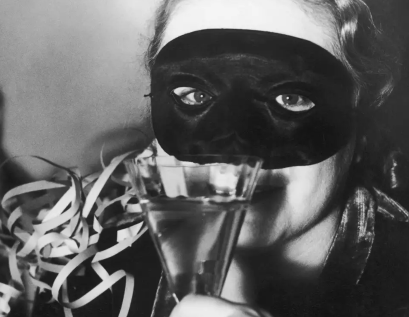 A person wearing a black eye mask and holding a glass, with festive streamers on their shoulder, looks directly at the camera. The image is in black and white.