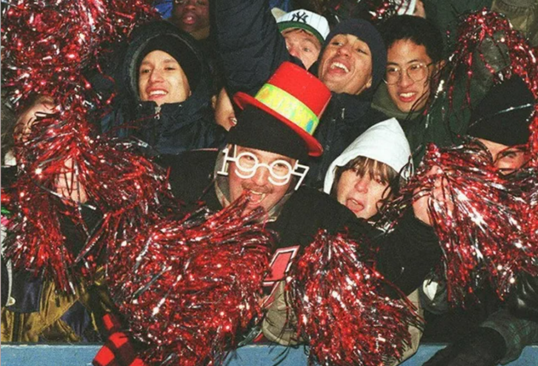 A crowd of excited people celebrate, some wearing hats and novelty glasses, and waving red pom-poms. One person wears large “2000” glasses and a black and red party hat, indicating a New Year celebration.