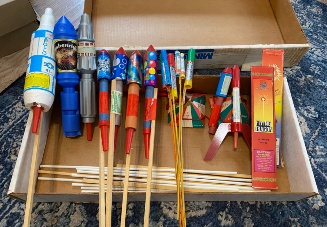 A cardboard box holds an assortment of colorful fireworks rockets and sparklers, arranged neatly on a blue patterned rug. Some rockets have labels and pointed cones, and several long wooden sticks are visible.