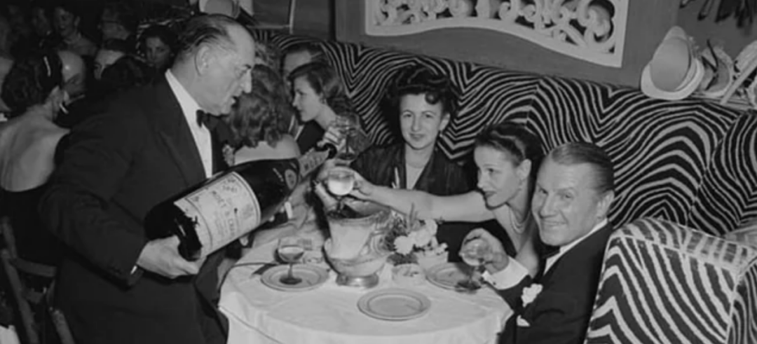 A waiter pours wine from a large bottle for elegantly dressed guests at a table in a lively restaurant with bold zebra-striped decor. The diners are smiling and engaged in conversation.