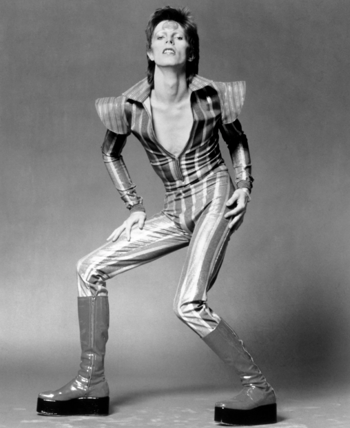 A person in a bold, striped, metallic jumpsuit with dramatic shoulder pads and platform boots strikes a dynamic pose against a plain backdrop, evoking a glam rock style.