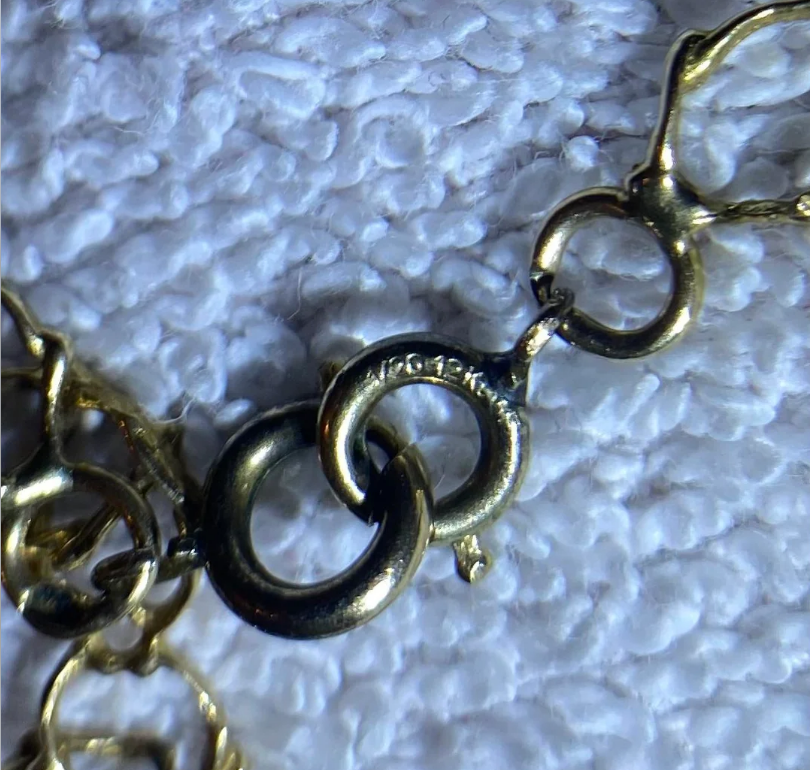Close-up of a gold chain necklace with circular links, placed on a textured white surface. One link is engraved with the word "VICTORIA.