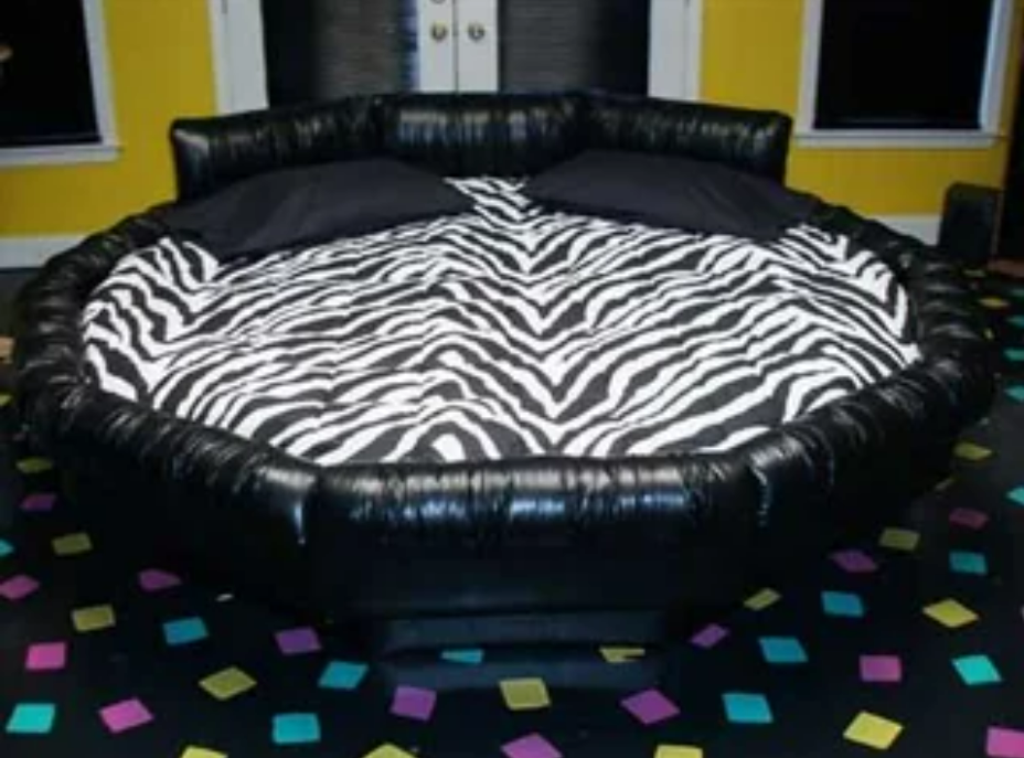 A round black leather bed with a zebra-print blanket and two black pillows sits in a room with yellow walls and colorful confetti-like squares on the floor.