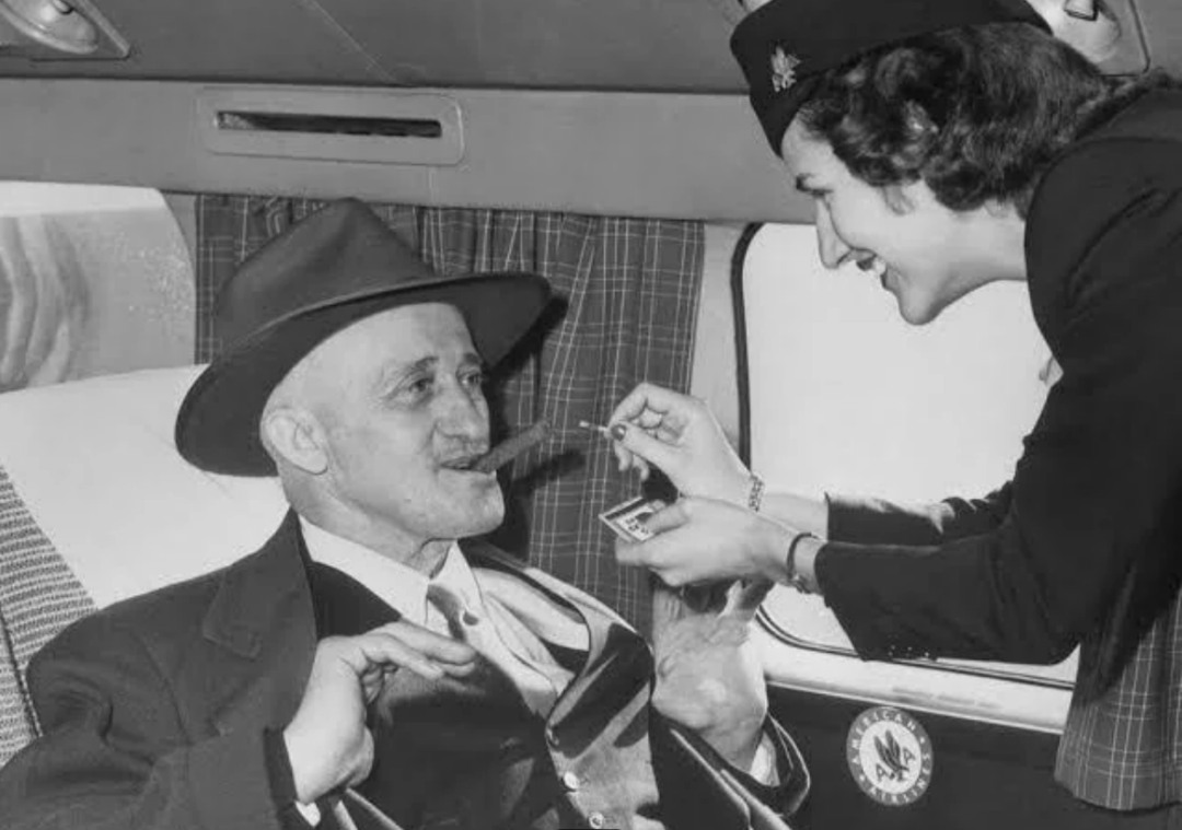 A smiling flight attendant lights a cigar for a seated man in a suit and hat inside an airplane. The man looks pleased, and the cabin interior features curtains and upholstered seats.