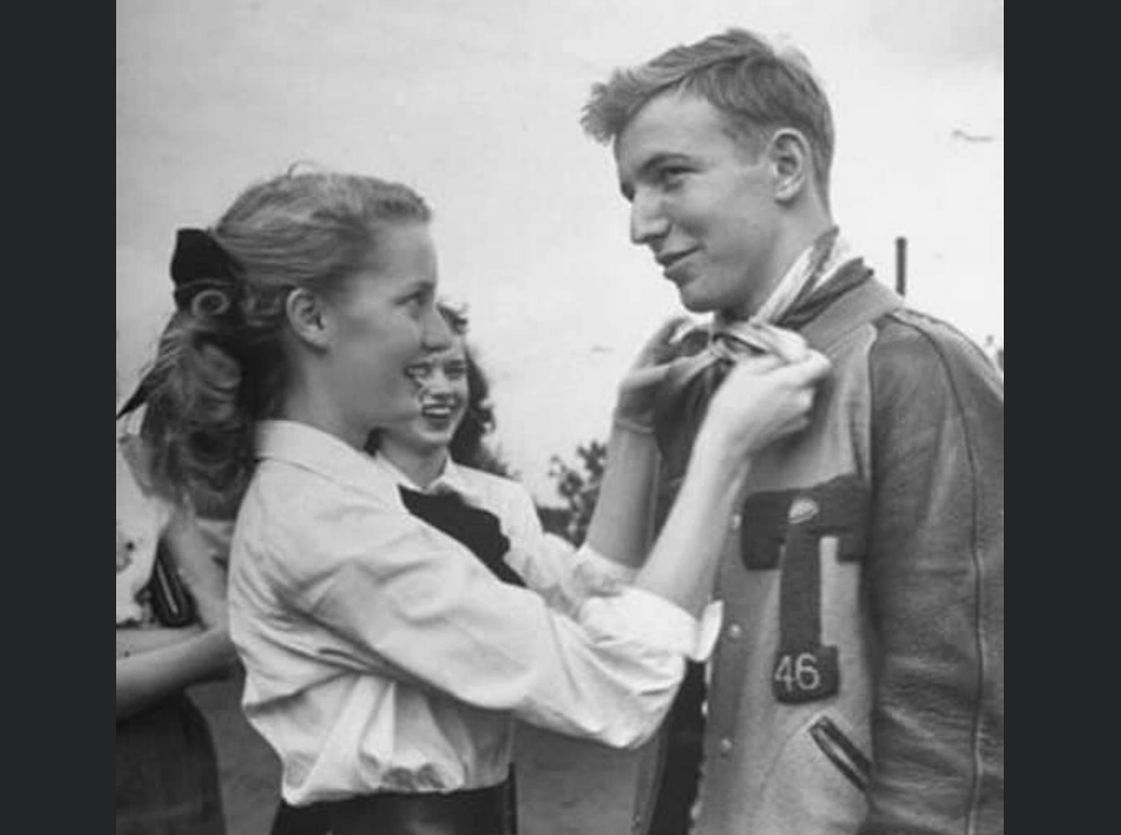 A young woman ties a scarf around a smiling young man's neck as another girl watches in the background. The man wears a letterman jacket, and they all appear happy outdoors in a vintage black-and-white photo.