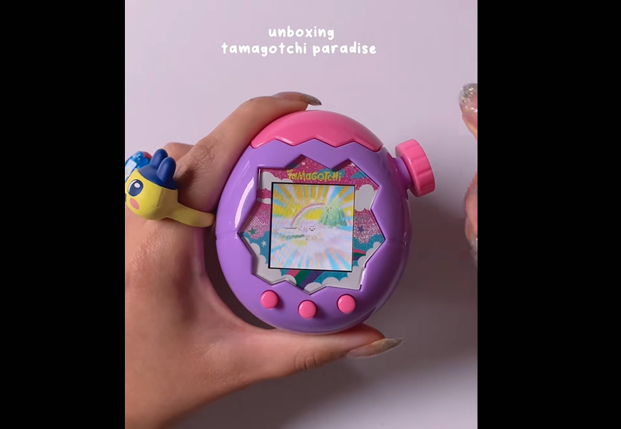 A person holds a pastel purple and pink Tamagotchi device with a digital screen and buttons. Their thumb wears a character-themed ring. The background text reads, "unboxing tamagotchi paradise.