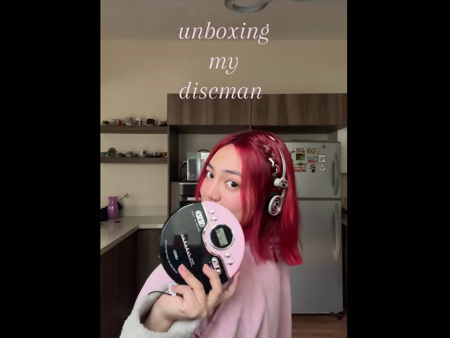 A person with bright red hair and headphones holds a black and pink Discman in a kitchen. Text above reads “unboxing my discman.” Shelves and a refrigerator are visible in the background.