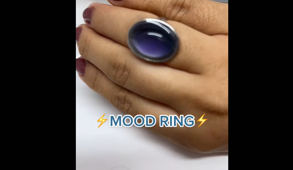 A hand with painted nails wears a large oval mood ring that is dark blue-purple. The words "MOOD RING" with lightning bolt emojis appear below the ring.