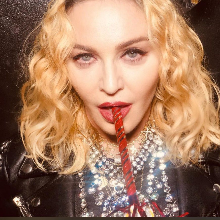 A woman with blonde wavy hair and bold makeup wears a black leather jacket and layered sparkling necklaces. She looks at the camera while holding a festive red party blower in her mouth.