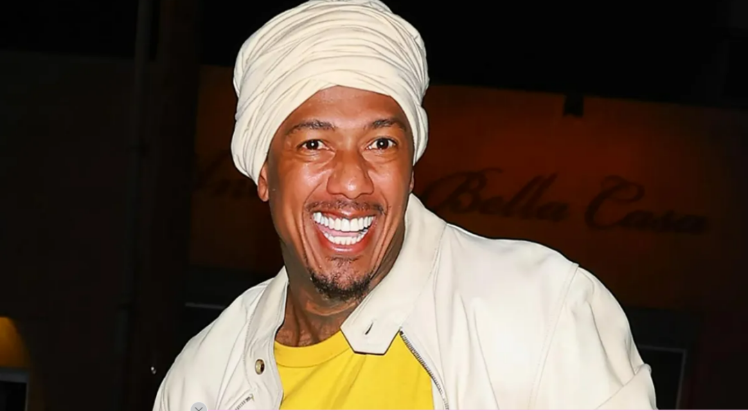 A man smiling widely, wearing a cream-colored turban, a matching jacket, and a yellow shirt, standing in front of a blurred, warmly lit background.