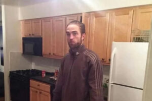 A man with light skin and brown hair pulled back stands in a kitchen with wooden cabinets, a white refrigerator, and a black stove. He is wearing a brown zip-up jacket and looking at the camera with a neutral expression.