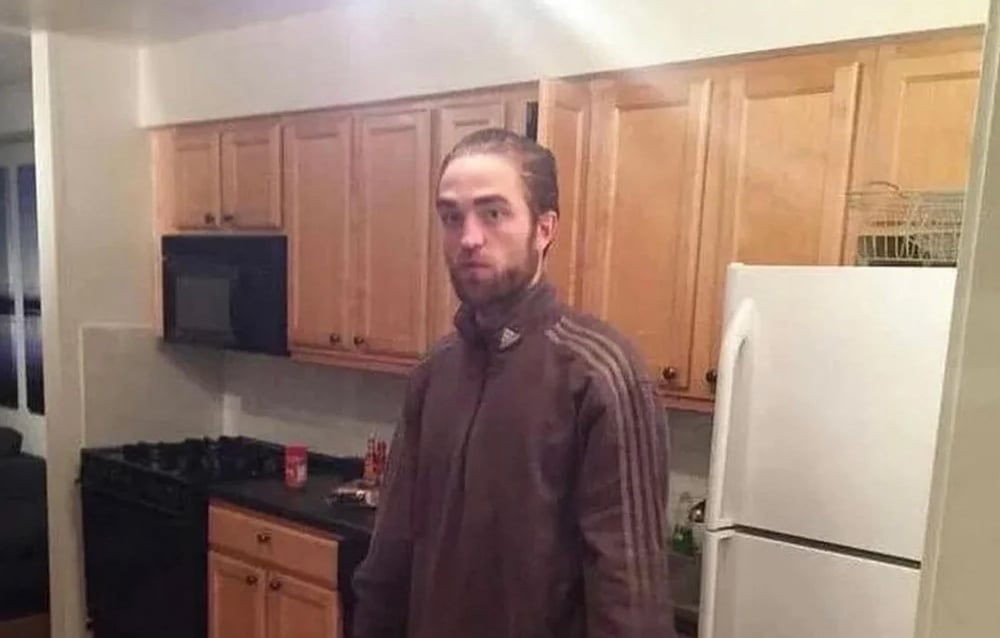 A man with light skin and brown hair pulled back stands in a kitchen with wooden cabinets, a white refrigerator, and a black stove. He is wearing a brown zip-up jacket and looking at the camera with a neutral expression.