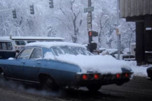 A blue vintage car with snow on its roof and trunk drives down a snowy, slushy street. Snow covers trees and the ground. Traffic lights and a building with a "JB" sign are visible in the background.
