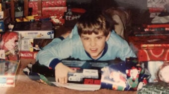 A young boy in blue pajamas lies on the floor, smiling excitedly while holding a Super Nintendo Entertainment System box among wrapped Christmas presents.