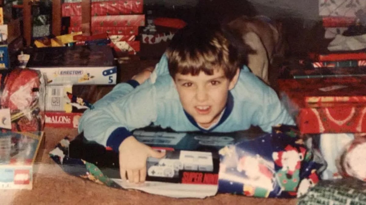 A young boy in blue pajamas lies on the floor, smiling excitedly while holding a Super Nintendo Entertainment System box among wrapped Christmas presents.