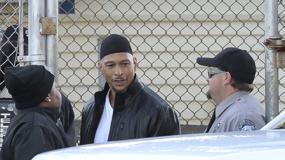 Three people talk near a chain-link fence. One man in a black jacket and cap stands between a woman in a black hat and a person in a uniform wearing a cap and sunglasses. A white vehicle is in the foreground.