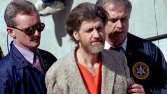 A bearded man in an orange shirt and gray coat is escorted by two law enforcement officers, one in a blue jacket with an official patch, outdoors in daylight.