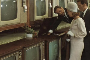 A man in a suit shows a console television to a woman and another man in a 1960s electronics store, with several vintage TVs displayed on shelves behind them.