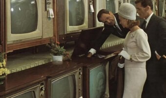 A man in a suit shows a console television to a woman and another man in a 1960s electronics store, with several vintage TVs displayed on shelves behind them.