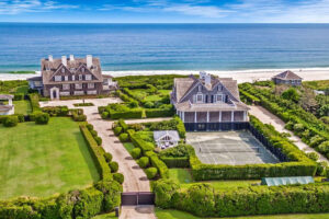 Aerial view of large beachfront estates with manicured lawns, tennis court, and direct access to a sandy beach; ocean waves and clear sky in the background.