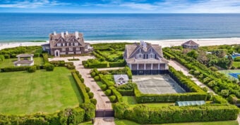 Aerial view of large beachfront estates with manicured lawns, tennis court, and direct access to a sandy beach; ocean waves and clear sky in the background.