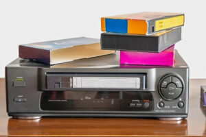 A black VHS player with buttons and a display sits on a wooden surface. Three VHS tapes in colorful cases are stacked on top of the player. The background is plain and white.