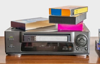 A black VHS player with buttons and a display sits on a wooden surface. Three VHS tapes in colorful cases are stacked on top of the player. The background is plain and white.
