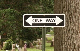 A &ldquo;One Way&rdquo; street sign with arrows pointing left and right stands in front of gravestones in a cemetery, creating a humorous contradiction.