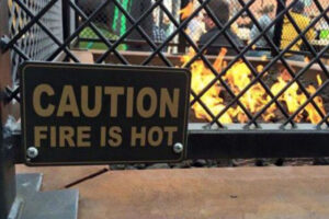 A metal sign on a fence reads "CAUTION FIRE IS HOT" in front of a fire pit with visible flames, with people gathered in the background.