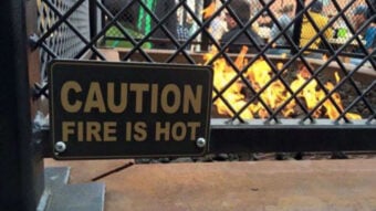 A metal sign on a fence reads "CAUTION FIRE IS HOT" in front of a fire pit with visible flames, with people gathered in the background.